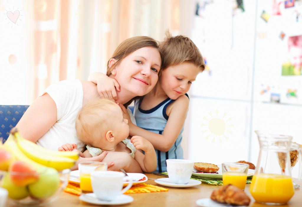 Mama Alltag erleichtern 5 Gewohnheiten Eine Mutter sitzt mit zwei kleinen Kindern an einem Frühstückstisch. Auf dem Tisch stehen Tassen, Gläser mit Orangensaft, Croissants, Obst und eine Karaffe. Die Szene wirkt warm und hell, mit Sonnenlicht, das durch die Vorhänge fällt, und einer gemütlichen Atmosphäre im Hintergrund mit einem Kühlschrank voller Fotos und Magneten.