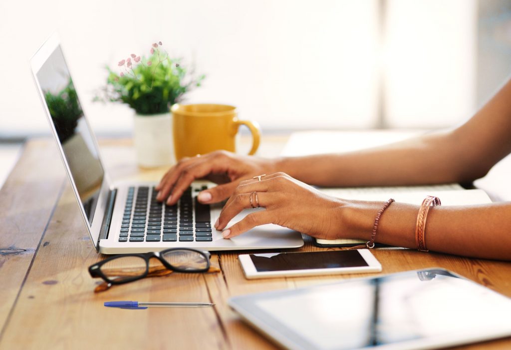 Eine Frau arbeitet konzentriert am Laptop auf einem hellen Holztisch. Neben ihr liegen Brille, Smartphone und Tablet, im Hintergrund stehen eine gelbe Tasse und eine kleine Pflanze. Das warme Licht und die ruhige Atmosphäre vermitteln Leichtigkeit und Fokus im Arbeitsalltag.